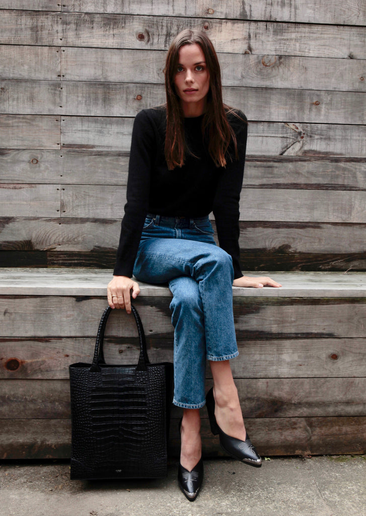 Model is holding the Duke Shopper tote bag in black croc embossed leather. 